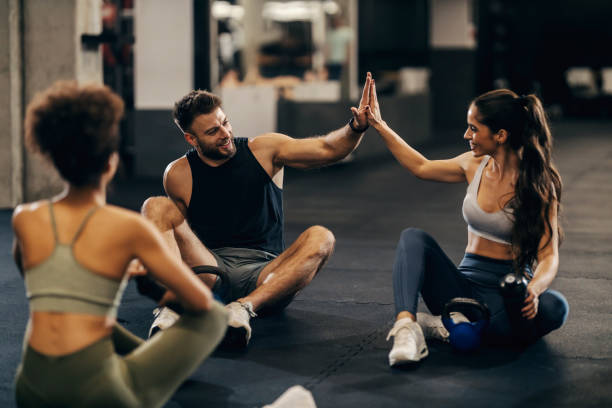 groupe de trois amis en forme qui se donnent des high five après l’exercice assis sur le sol d’un gymnase. - salle de sport photos et images de collection